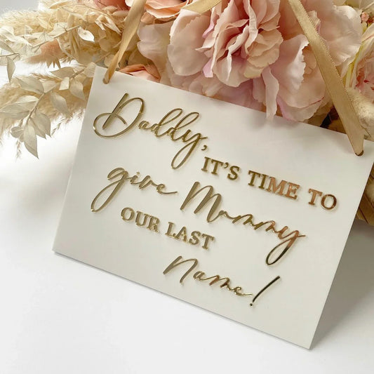 Wedding proposal sign with gold script on white plaque, surrounded by blush pink flowers.